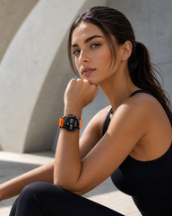 Woman wearing a smartwatch with an orange band, sitting against a concrete wall.